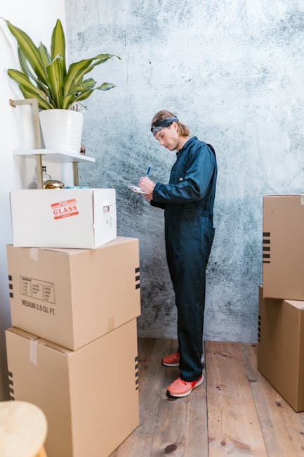 A woman with curly brown hair, dressed in a white t-shirt and light-colored pants, is crouching on a concrete floor inside a room, surrounded by several large cardboard boxes used for packing. She is