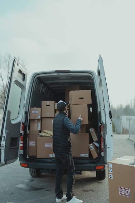 A man wearing a dark jacket, jeans, and a black baseball cap is loading or preparing to load multiple cardboard boxes into the rear of a white panel van parked outdoors. The boxes vary in size, with some stacked inside the van and some positioned near the open rear doors, which reveal the vehicle's spacious interior. The van is situated on a paved surface, possibly a driveway or parking area, with a background of trees and overcast sky. The scene depicts the process of house relocation or furniture transport, with Man with Van Monken Hadley involved in a home removal service that includes packing, loading, and transport logistics.