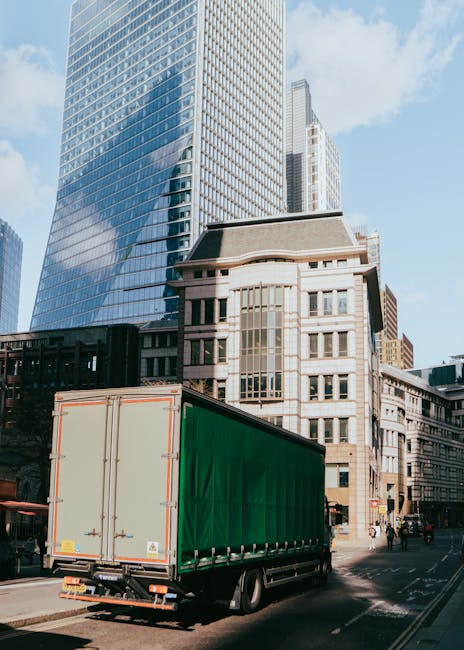 A large delivery truck with a green cargo area is parked on the street in an urban area, in front of modern glass skyscrapers and older stone buildings. The truck is positioned near the curb, with its rear doors closed, ready for loading or unloading. The scene shows the loading process typical of home relocation or furniture transport, with no visible moving personnel or items being moved at this moment. The street appears busy with pedestrians and urban activity, with natural daylight illuminating the scene. Man with Van Monken Hadley, a professional removals company, frequently uses such vehicles for house removals, ensuring secure transport of furniture and boxes during relocation projects.