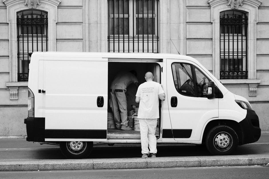 A man wearing a dark jacket, jeans, and a black baseball cap is loading or preparing to load multiple cardboard boxes into the rear of a white panel van parked outdoors. The boxes vary in size, with some stacked inside the van and some positioned near the open rear doors, which reveal the vehicle's spacious interior. The van is situated on a paved surface, possibly a driveway or parking area, with a background of trees and overcast sky. The scene depicts the process of house relocation or furniture transport, with Man with Van Monken Hadley involved in a home removal service that includes packing, loading, and transport logistics.