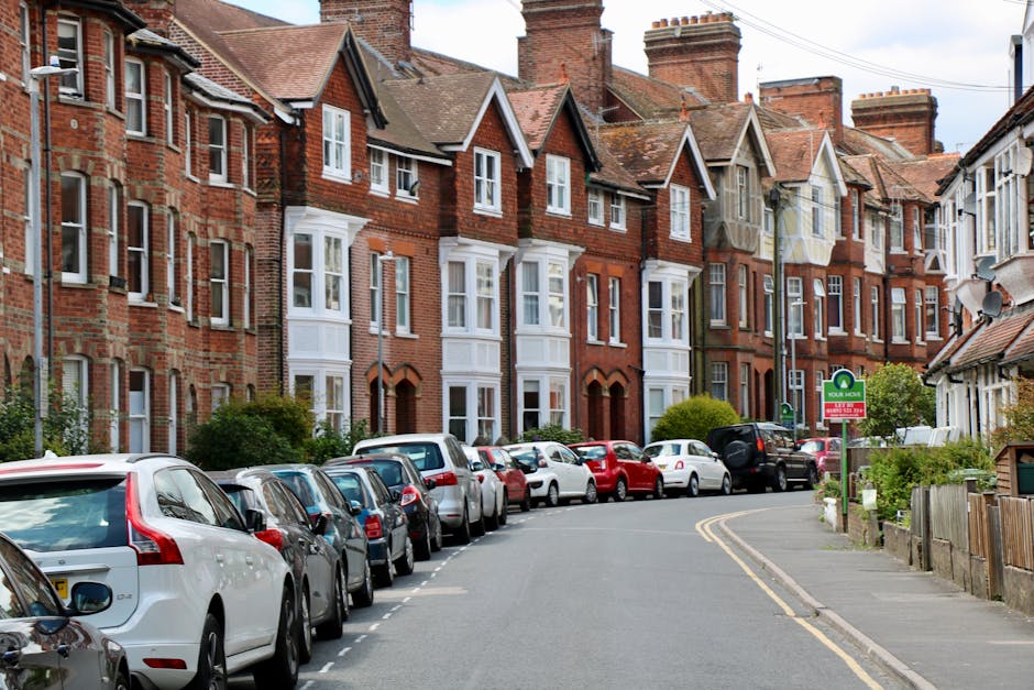 A residential street lined with multiple three-storey brick houses featuring white window frames and pitched roofs, situated behind a row of parked cars including sedans, hatchbacks, and SUVs on the curbside. The street has a slight bend towards the background, with a narrow sidewalk on the right side, bordered by a low wooden fence and a small garden area with bushes and greenery. A green sign indicating a resident parking zone is visible on a lamppost, and the sky above is partly cloudy. The scene depicts an urban environment suitable for house removals and furniture transport, with a focus on the typical setting for home relocation process, including the loading and parking logistics managed by local services such as Man with Van Monken Hadley.