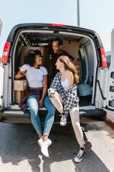Two young women are sitting inside the open rear of a white moving van, engaging in conversation and smiling, with one gesturing animatedly. The woman on the left has curly black hair, is wearing a li