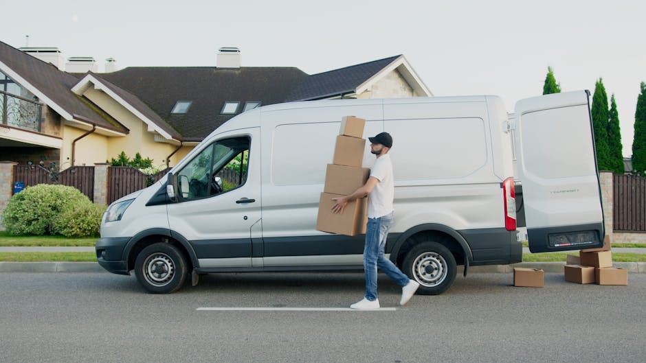 Two young women are sitting inside the open rear of a white moving van, engaging in conversation and smiling, with one gesturing animatedly. The woman on the left has curly black hair, is wearing a li
