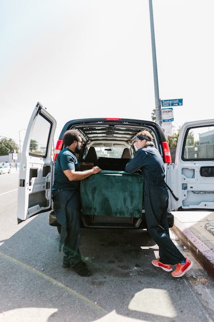 Two movers from Man with Van Monken Hadley, dressed in navy uniforms with protective eyewear, are engaged in a home relocation process, loading a large, dark green wrapped piece of furniture into the open cargo area of a black panel van on a paved street. The vehicle's rear doors are fully open, revealing the clean interior with elements of packaging materials such as foam padding and blankets used for securing furniture during transport. One mover is holding the furniture steady while the other adjusts or guides it, demonstrating careful handling during the furniture transport phase. The surrounding environment includes a street sign for Somerset Close, some nearby parked vehicles, and a partly cloudy sky, indicating daytime conditions. This loading process is part of a professional removals service, emphasizing safe packing, loading, and transportation of household belongings, consistent with the company's offerings in house removals and urgent same-day move solutions.