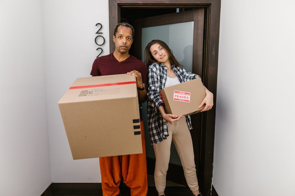 A man and a woman stand inside the entrance of a residential property, holding cardboard boxes related to house removals. The man, dressed in an orange pair of trousers and a maroon t-shirt, is holding a large moving box with packing tape and labels, positioned in front of him. The woman, wearing beige pants and a plaid shirt over a white top, is holding a smaller box marked with a 'Fragile' sticker, and leans slightly against the doorframe. The background shows a dark wooden door, with the door number visible, leading to the interior of the home. The scene is well-lit, possibly with natural daylight, highlighting the furniture, packing materials, and the involved moving process. The environment indicates a home relocation setup, with emphasis on furniture transport, packing, and loading activities that are typical of emergency same-day removals, as provided by Man with Van Monken Hadley.