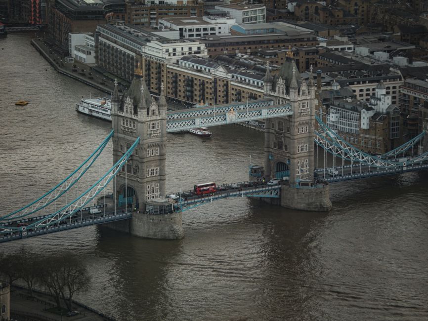 An aerial view of Tower Bridge in London spanning the River Thames, with its distinctive twin towers and blue suspension cables visible. The bridge is partially open, showing a red double-decker bus and vehicles on the roadway. Surrounding the bridge are modern and historic buildings in the City of London, with some boats navigating the river. The scene is captured in natural daylight, with overcast weather creating a diffuse lighting effect. This image relates to house removals and furniture transport, illustrating the typical urban environment where local moving services like those offered by Man with Van Monken Hadley operate, often involving logistical planning around London's iconic landmarks and bridge crossings during home relocation processes.