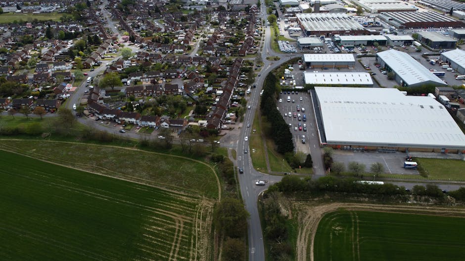 Aerial view of a residential neighbourhood with closely spaced houses and narrow streets alongside commercial warehouses and industrial buildings, all surrounded by green fields and trees. Vehicles are seen on the roads, and the area includes a mix of urban and semi-rural elements. The scene shows a typical home removal logistics environment, with some cars parked near the houses and industrial units, as well as open spaces suitable for loading and unloading goods. The lighting suggests daytime conditions, and the image reflects a typical setting where house removals and furniture transport might take place, managed by companies like Man with Van Monken Hadley.