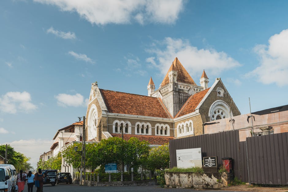 The image shows a historic church with a prominent pitched roof covered in red tiles, large arched windows, and a tall central tower topped with a pointed spire. The facade features decorative stonework and Romanesque-style round arches, set against a bright blue sky with scattered clouds. In the foreground, several people are walking along a pavement lined with trimmed green trees, with parked cars adjacent to the street. To the right, a dark-colored fence partially encloses an area near the church, with a sign and a red mailbox visible on a low stone wall. The scene appears during daylight, with soft natural lighting highlighting the church's architectural details. This setting reflects a typical environment where house removals or furniture transport may take place, with the church as a notable landmark nearby. The image is relevant to home relocation and moving services, which could include logistics around parking and access for a professional removal company like Man with Van Monken Hadley.