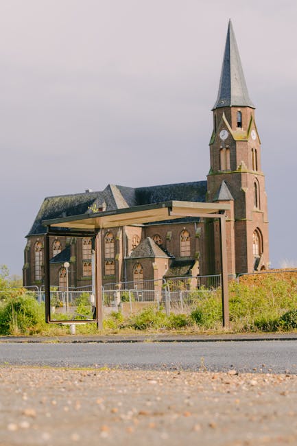 A row of parked cars lining a narrow street leading to a historic church with a tall, pointed spire and a large clock on its brick facade. The church is surrounded by lush green trees, and the scene is captured in natural daylight. In the foreground, a man with a van, which is likely associated with Man with Van Monken Hadley, is engaged in a home relocation or furniture transport operation, with cardboard boxes, wrapped furniture, and moving blankets visible near the vehicle. The loading process appears to be taking place close to the church entrance, with some items on dollies or being carried by movers in preparation for the move. The setting suggests a professional packing and moving service focused on house removals near All Saints' Church, Monken Hadley, highlighting the logistical aspects of furniture transport and home relocation within a residential or urban environment.