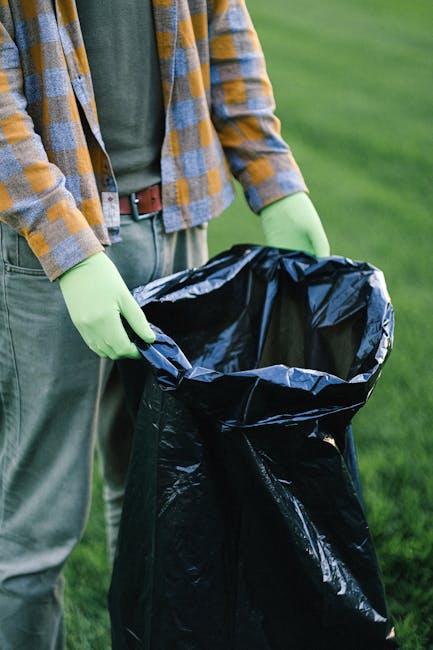 A person wearing a plaid shirt, green gloves, and jeans is standing outdoors on a grassy area, holding a black trash bag open in both hands, preparing for waste disposal or rubbish collection. The scene is well-lit with natural daylight, and the background consists of blurred green grass and possibly trees. This image illustrates the cleaning or waste management process associated with home relocation or packing and moving services, relevant to the scope of house removals offered by Man with Van Monken Hadley. The focus is on the action of handling waste materials in preparation for removal, supporting efficient packing, or clearing before a move.