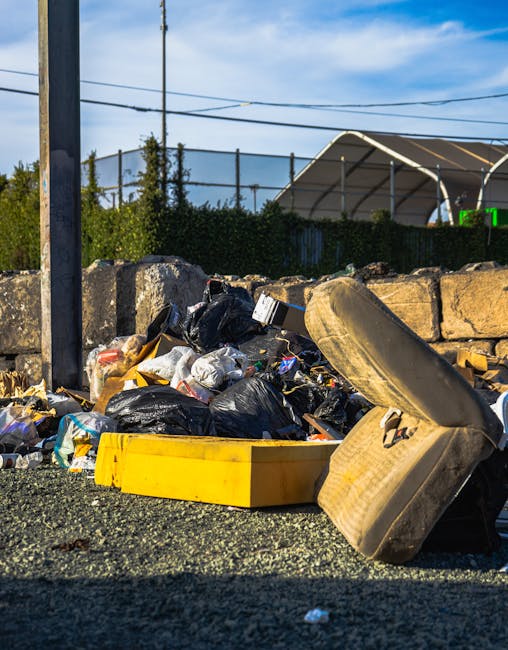 A pile of mixed household waste including black plastic bin bags, cardboard boxes, and discarded items such as an old, dirty beige upholstered armchair with visible stains and fabric tearing, all placed on a gravel surface outside a property. The waste is stacked haphazardly against a stone wall, with some items partially spilling onto the ground. In the background, there is a metal fence, utility poles with overhead wires, and a clear blue sky with a few scattered clouds. The scene appears to be part of a home clearance or waste disposal process, with an emphasis on visible packaging materials and furniture ready for removal, aligning with professional local removals and clearance services provided by Man with Van Monken Hadley.