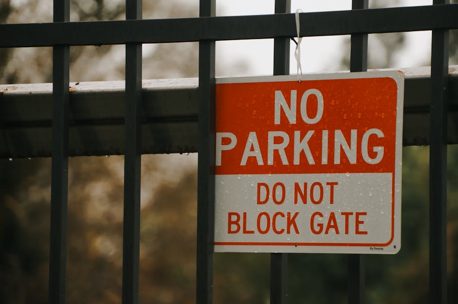 A rectangular metal sign with a red and white background mounted on a black metal fence. The sign displays the text 'NO PARKING' in large white capital letters at the top, followed by 'DO NOT BLOCK GATE' in smaller red capital letters beneath. The fence has vertical bars and runs horizontally across the image, with a blurred outdoor background showing autumnal trees and foliage. The sign is positioned in an outdoor setting, possibly near a residential property or commercial premises in Monken Hadley, indicating restrictions on parking in the vicinity of a gate. This scene relates to moving and home relocation contexts where parking limitations can affect the loading and unloading process, and the sign is a typical part of relocation logistics managed by companies like Man with Van Monken Hadley.