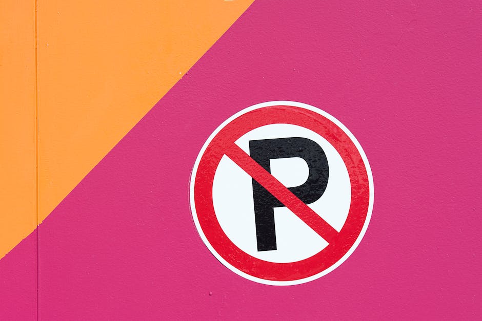 A circular 'No Parking' sign with a bold black letter 'P' crossed out in red, mounted on a pink wall with a diagonal section of orange paint in the top left corner. The sign is positioned near a residential property, indicating parking restrictions likely affecting the home relocation or furniture transport process. This image relates to house removals and moving services offered by Man with Van Monken Hadley, highlighting the importance of planning for parking limitations during a house move or furniture transport, especially when space is restricted or parking is not permitted at the property or along the route to the driveway or loading area.