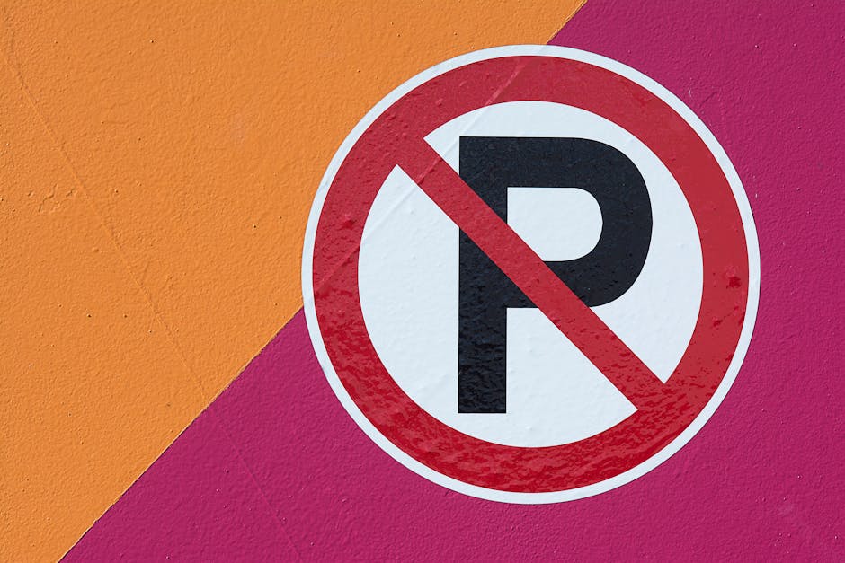 A rectangular metal sign with a red and white background mounted on a black metal fence. The sign displays the text 'NO PARKING' in large white capital letters at the top, followed by 'DO NOT BLOCK GATE' in smaller red capital letters beneath. The fence has vertical bars and runs horizontally across the image, with a blurred outdoor background showing autumnal trees and foliage. The sign is positioned in an outdoor setting, possibly near a residential property or commercial premises in Monken Hadley, indicating restrictions on parking in the vicinity of a gate. This scene relates to moving and home relocation contexts where parking limitations can affect the loading and unloading process, and the sign is a typical part of relocation logistics managed by companies like Man with Van Monken Hadley.
